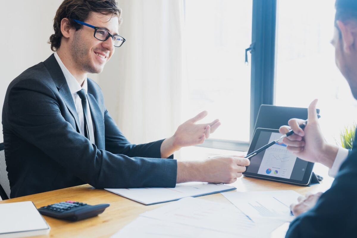 Two businessmen in formal attire discussing a business report in an office setting with a tablet.